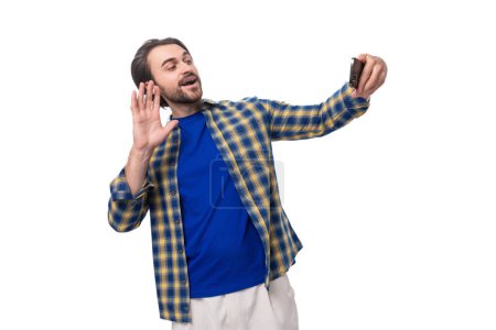 Foto de Un joven caucásico brutal hombre con una cara sin afeitar vestido con una camisa a cuadros azul habla a través de enlace de vídeo smartphone. - Imagen libre de derechos