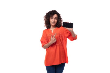 Téléchargez les photos : Jeune élégante femme brune caucasienne frisée vêtue d'un chemisier orange montre écran de téléphone. concept publicitaire. - en image libre de droit