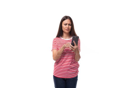 Foto de Una mujer caucásica bastante joven con pelo liso oscuro en gafas y una camiseta roja a rayas utiliza aparatos modernos como un teléfono inteligente. - Imagen libre de derechos