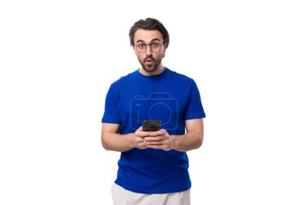 Foto de Un europeo de 30 años de edad, con el pelo negro y una barba en una camiseta azul utiliza un teléfono inteligente y mira a la cámara con sorpresa. - Imagen libre de derechos