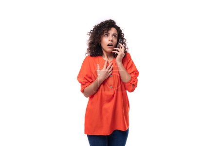 Téléchargez les photos : Jeune femme européenne aux cheveux noirs bouclés dans un chemisier orange parle au téléphone. - en image libre de droit