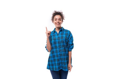 Foto de Joven mujer europea de cabello oscuro vestida informalmente con una camisa a cuadros azul se inspira y señala con el dedo hacia arriba. - Imagen libre de derechos
