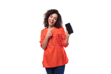 Téléchargez les photos : Jeune femme brune caucasienne aux cheveux bouclés habillé en chemisier orange points smartphone avec maquette d'écran. - en image libre de droit