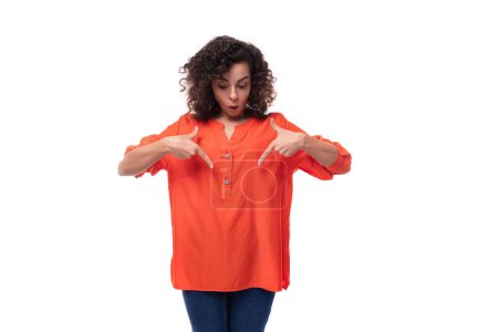 Téléchargez les photos : Portrait d'une jeune femme brune caucasienne aux cheveux bouclés habillée d'une chemise orange pointant sa main en surprise. - en image libre de droit