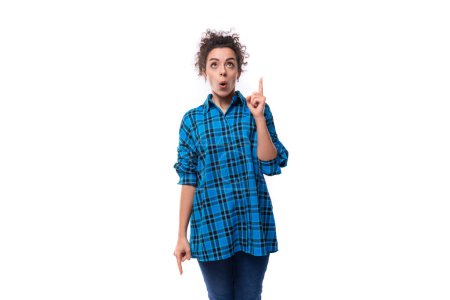 Téléchargez les photos : Jeune femme européenne aux cheveux bruns vêtue informellement d'une chemise à carreaux bleue est inspirée et pointe son doigt vers le haut. - en image libre de droit