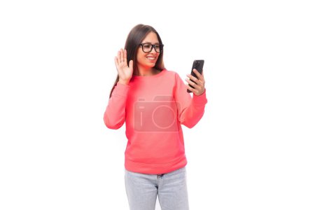 Téléchargez les photos : Jolie jeune femme brune caucasienne aux cheveux raides vêtue d'un sweat-shirt rose et d'un jean bavardant par appel vidéo sur un smartphone sur fond blanc avec espace de copie. - en image libre de droit