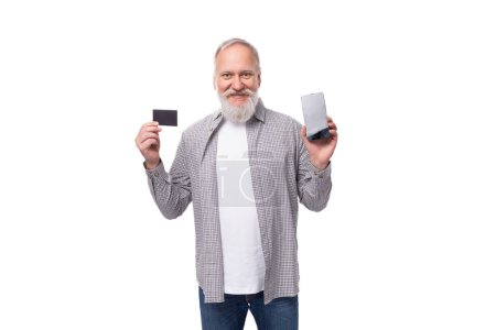Téléchargez les photos : Un bel homme pensionné avec une barbe et une grosse moustache vêtue d'une chemise et d'un jean tient une maquette de carte en plastique et un smartphone avec l'écran tourné vers l'avant. Concept de commerce électronique. - en image libre de droit