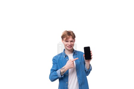 Foto de Joven encantador europeo con el pelo dorado vestido con una camisa de mezclilla azul sostiene un teléfono inteligente en su mano. - Imagen libre de derechos
