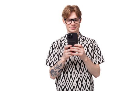 Téléchargez les photos : Jeune homme caucasien beau avec des cheveux rougeâtres dorés dans une chemise d'été noir et blanc prend une photo sur un smartphone. - en image libre de droit