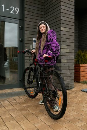 Eine hübsche junge Frau mit Piercings und Dreadlocks im lila Pelzmantel fährt Fahrrad.