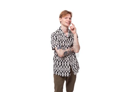 Foto de Retrato de un joven informal pensante con el pelo rojo y un tatuaje en los brazos, vestido con una camisa de manga corta con un patrón. - Imagen libre de derechos