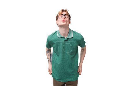 Foto de Joven jengibre brillante con un tatuaje en el brazo vestido con una camiseta verde de manga corta envía un beso de aire. - Imagen libre de derechos
