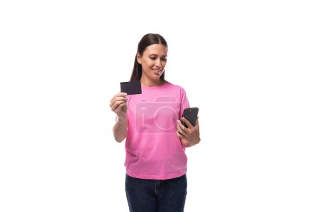 Foto de Joven mujer morena delgada en camiseta básica rosa con tarjeta de crédito maqueta y teléfono inteligente sobre fondo blanco con espacio para copiar. - Imagen libre de derechos