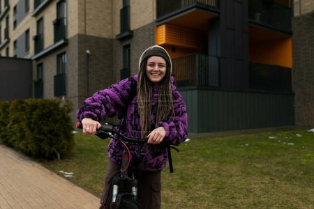 Jeune femme mignonne avec piercing et dreadlocks en vélo dans un environnement urbain.