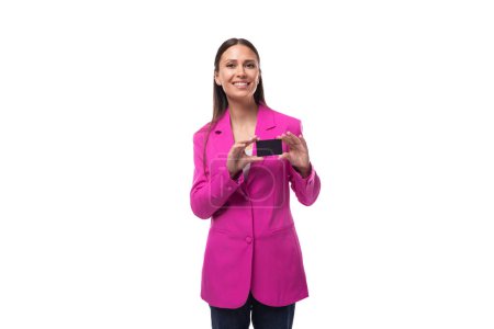 junge fröhliche positive Büroangestellte mit schwarzen Haaren in fliederfarbener Jacke demonstriert eine Kreditkarte mit einer Attrappe.