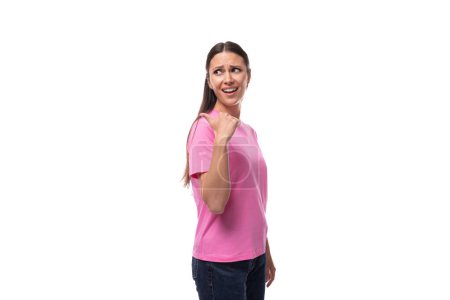 Téléchargez les photos : Jeune femme brune dans un t-shirt rose pointe sa main sur le côté sur un fond blanc avec espace de copie. - en image libre de droit