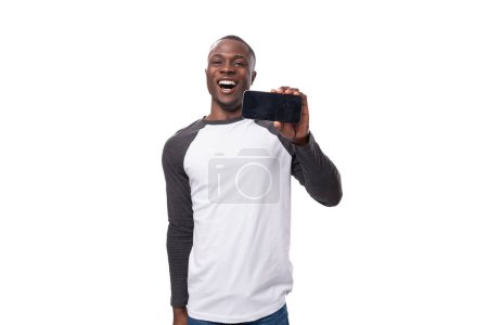 Foto de Joven americano con corte de pelo corto vestido con un suéter de manga larga informa de la noticia señalando con su mano a un teléfono inteligente con una maqueta para la publicidad. - Imagen libre de derechos