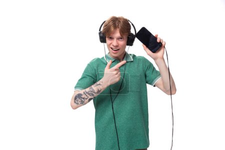 Foto de Joven alegre estudiante europeo delgado con el pelo rojo en una camiseta verde escucha música fuerte en los auriculares. - Imagen libre de derechos