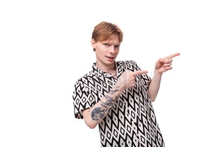 Foto de Un joven guapo con el pelo rojo y un tatuaje en los brazos, vestido con una camisa de manga corta, señala con la mano el espacio para la publicidad. concepto de negocio. - Imagen libre de derechos