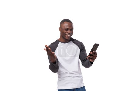 Foto de Un joven africano con un corte de pelo corto vestido con un suéter de manga larga de verano utiliza transferencia en línea usando un teléfono inteligente. - Imagen libre de derechos