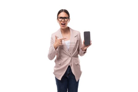 jeune brune intelligente à queue de cheval vêtue d'une veste légère montre un écran de smartphone avec une maquette.