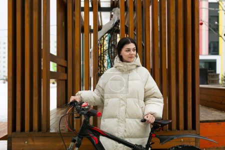 Una mujer está de pie junto a una bicicleta estacionada frente a una estructura de madera. Ella parece estar tomando un descanso o admirando los alrededores en un día soleado.