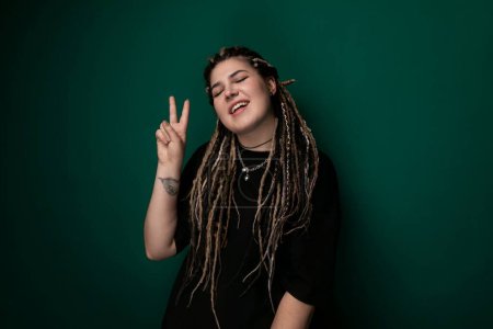 A woman with shoulder-length dreadlocks is seen in the image, raising her right hand to make a peace sign gesture. She is wearing a casual outfit and standing against a plain background.
