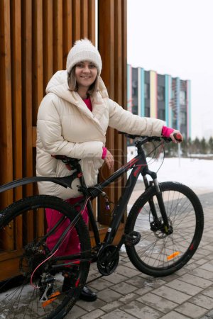 Eine Frau steht neben einem Fahrrad auf einem Gehweg. Sie scheint sich auf das Radfahren vorzubereiten, mit Gebäuden im Hintergrund.