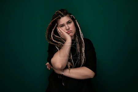 A woman with dreadlocks is standing confidently in front of a vivid green background. Her hair is neatly styled into long, thin dreadlocks, and she is dressed in casual attire. The green backdrop