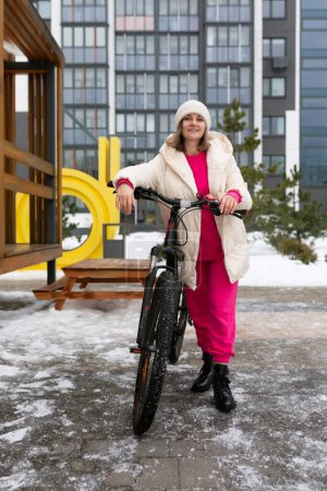 A woman is standing next to a bicycle on a snowy day. She is dressed warmly, with snowflakes falling around her. The bike is covered in a light dusting of snow, contrasting with the white landscape.