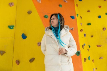 A woman is standing in front of a climbing wall, wearing climbing gear. She appears to be assessing the wall before attempting to climb. The wall is colorful and has various holds and routes marked