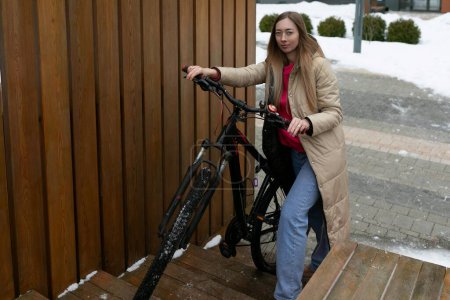 A woman is standing beside a bicycle in a snowy landscape. She is rugged up in warm clothing, looking at the camera with a neutral expression. The bike is covered with a dusting of snow, highlighting