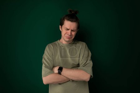A man is standing with his arms crossed in front of a bright green wall, with a neutral expression on his face. The wall serves as a simple backdrop for the mans pose.