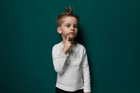A young boy is standing alone against a vibrant green wall, looking straight ahead. He is wearing casual clothes and appears to be deep in thought.