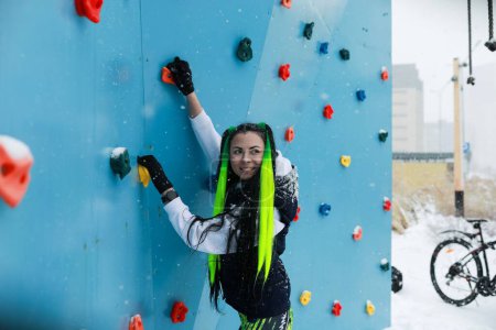 A woman with bright green hair stands confidently next to a colorful climbing wall, ready to tackle the challenging route in front of her.