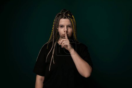 A woman with dreadlocks is raising her index finger to her lips, signaling for silence or secrecy. Her facial expression appears serious as she gestures to maintain quiet.