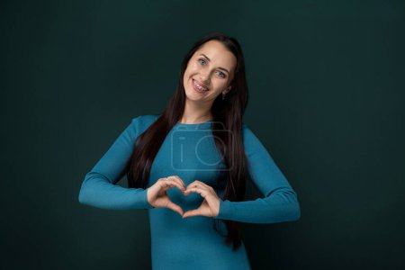 A woman is forming a heart shape with her hands, symbolizing love and affection. She stands against a plain background, focusing on the hand gesture and its significance.