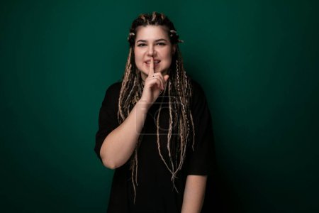 Une femme aux longues dreadlocks se tient devant un mur vert. Elle semble confiante et élégante, mettant en valeur sa coiffure unique.