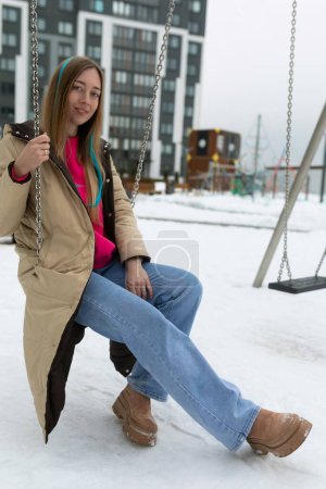 Téléchargez les photos : Une femme portant des vêtements d'hiver est assise sur une balançoire couverte de neige. Les flocons de neige tombent doucement autour d'elle, créant une scène d'hiver sereine. - en image libre de droit