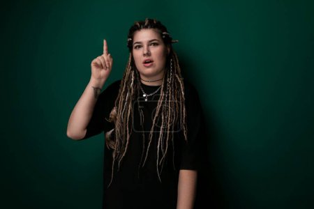 A woman with dreadlocks is pointing at an object or direction off-camera, her hand raised and finger extended. She appears engaged and focused on the subject of her gesture.