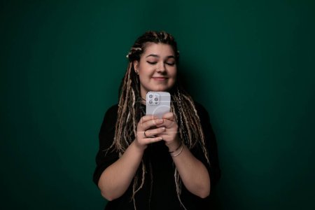 A woman with dreadlocks is seen holding a cell phone in her hand. She appears focused on the screen, possibly texting or browsing. Her dreadlocks are neatly styled and frame her face. The background