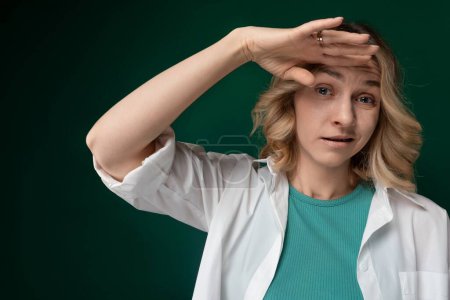 Une femme portant une chemise verte est montrée avec les mains posées sur sa tête dans un geste de détresse ou de frustration. Elle semble exprimer de fortes émotions à travers son langage corporel.