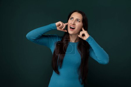 Una mujer que lleva una camisa azul se muestra sosteniendo sus manos a sus oídos en un gesto de escuchar o cubrirlos debido a un fuerte ruido o malestar.