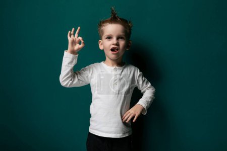 Ein kleines männliches Kind steht vor einer soliden grünen Wand und blickt geradeaus. Der Junge wirkt neugierig und aufmerksam, seine Hände ruhen an seinen Seiten..