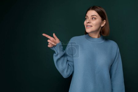 Une femme se tient debout devant un mur vert solide. Elle semble calme et composée, regardant directement la caméra. Le fond est minimaliste, attirant l'attention sur la présence des femmes.