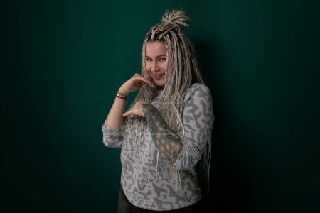 A woman with dreadlocks is standing in front of a vibrant green wall. She is looking directly at the camera with a confident expression. The texture of her hair contrasts with the smooth surface of