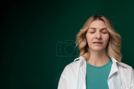 A woman with her eyes closed is seen standing in front of a solid green background. Her posture is upright, and she appears to be deep in thought or meditation.