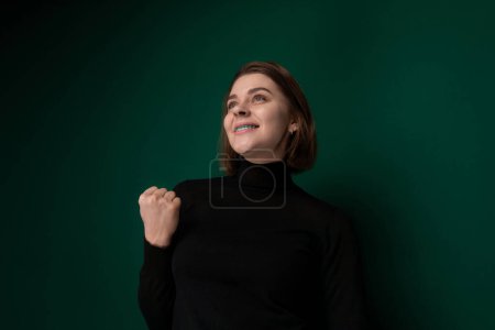 A woman with short brown hair, wearing a black turtleneck, is captured mid-smile looking up and to the side. Her expression appears cheerful and confident, set against a rich, dark green backdrop that