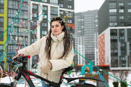 Eine Frau in Winterkleidung steht neben einem Fahrrad in einer verschneiten Szene.