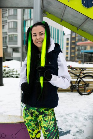 A woman with neon green hair standing in a snowy landscape. She is dressed warmly and looking around, surrounded by white snow.